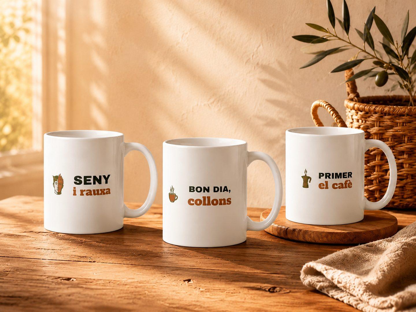 Tres tasses de ceràmica blanca amb els dissenys «Seny i rauxa», «Bon dia, collons» i «Primer el cafè» sobre una taula de fusta